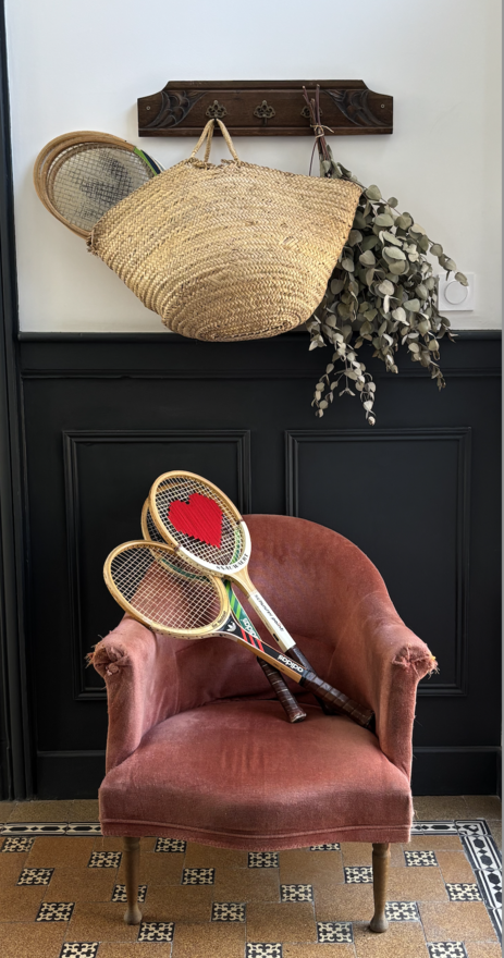 Capture d’écran 2026-07-13 à 10.20.22 Fauteuil vintage en velours terracotta décoré de deux raquettes de tennis en bois anciennes, dont une personnalisée avec un cœur rouge, sous un porte-manteau en bois avec un panier en osier, une raquette vintage et des branches d'eucalyptus.